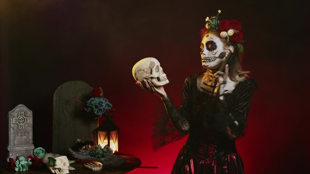 Santa muerte model on phone call holding skull, talking on smartphone line and acting creepy in studio. Answering telephone for remote chat, wearing holy mexican goddess costume.