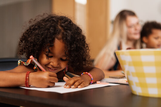 Little Girl Sitting At The Table And Writing On A Piece Of Paper
