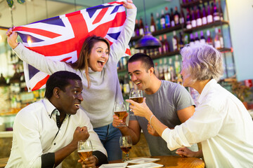 Happy multiracial friends holding the flag of England, rejoicing winning football game and drinking...
