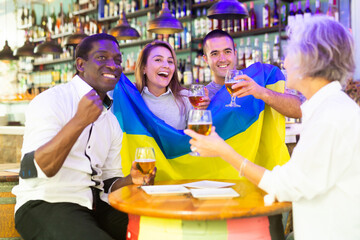 Cheerful diverse soccer supporters holding the flag of Ukraine and enjoing beer, spending time together in the sport bar
