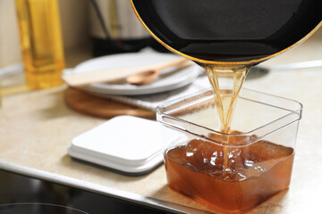 Pouring used cooking oil from frying pan into container on beige table in kitchen, closeup