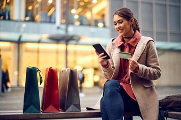 Happy woman using credit card and cell phone while enjoying in shopping day in city.