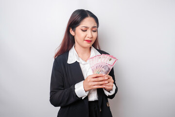 A confused young businesswoman is wearing black suit and holding cash money in Indonesian rupiah...