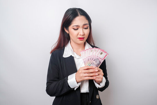 A confused young businesswoman is wearing black suit and holding cash money in Indonesian rupiah isolated by white background