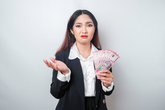 A confused young businesswoman is wearing black suit and holding cash money in Indonesian rupiah isolated by white background