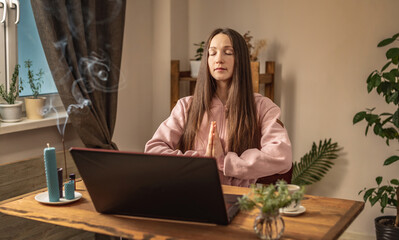 Woman is sitting at a table in front of a laptop, meditating and relaxing while listening to an...
