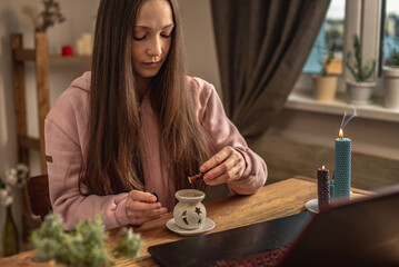 Woman sits in front of a laptop, listens to an instructor or a video lecture on meditation and relaxation and drips essential oil into an aroma lamp. Concept of a remote therapy and meditation session