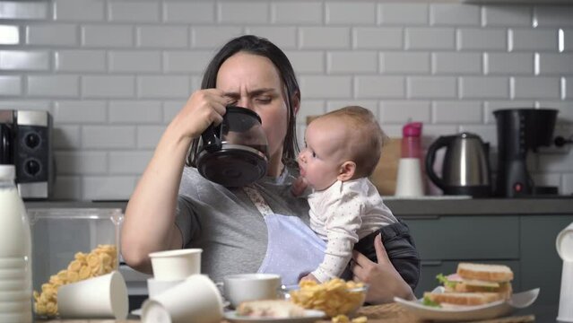 Tired woman drinking coffee and trying to eat while holding her baby in hands in messy kitchen. Single mother and child caring concept