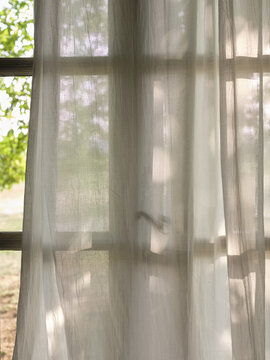 View through linen curtains and wooden window of green trees in garden