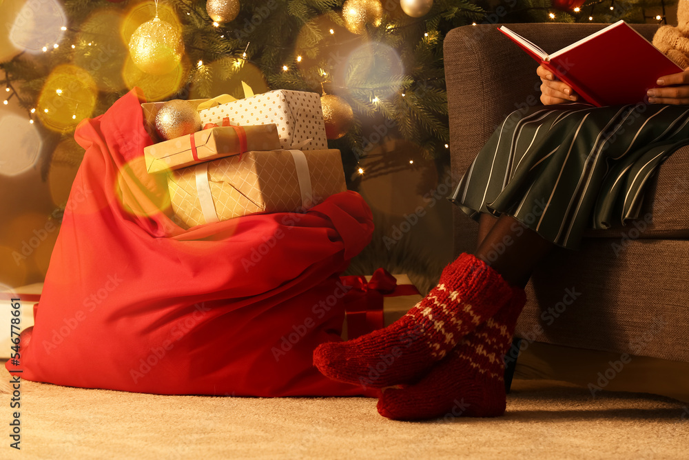 Wall mural woman reading book while sitting in armchair near santa bag full of gifts