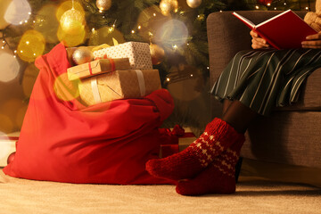 Woman reading book while sitting in armchair near Santa bag full of gifts