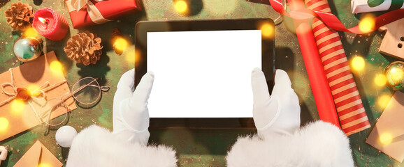 Hands of Santa Claus with tablet computer on green background with Christmas decor and gifts, top...