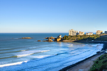 back view of biarritz beach on a sunny autumn morning. The sun hits the white buildings of the beach on the cliffs, below the beach the promenade in the shade, the waves break calmly against the sand 
