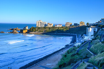 back view of biarritz beach on a sunny autumn morning. The sun hits the white buildings of the beach on the cliffs, below the beach the promenade in the shade, the waves break calmly against the sand 