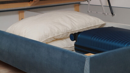 Storage drawer under bed with blue suitcase and white pillows indoors, closeup