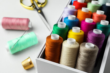 Box with colorful sewing threads, scissors and thimble on white marble table, closeup