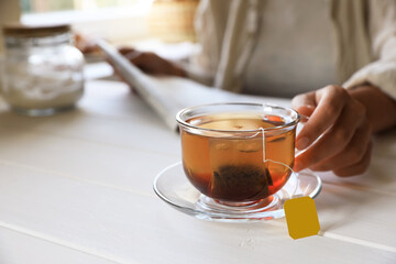 Woman holding glass cup with tea bag at white wooden table, closeup. Space for text