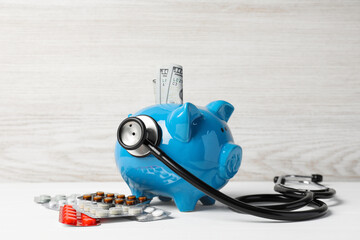 Light blue ceramic piggy bank with money, stethoscope and pills on white wooden table. Medical insurance