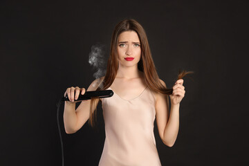 Upset young woman with flattening iron on black background. Hair damage