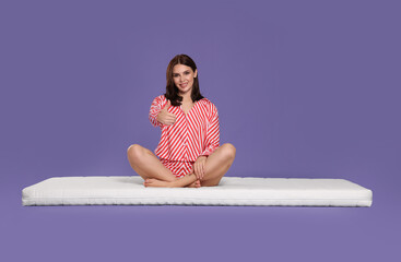 Obraz na płótnie Canvas Young woman sitting on soft mattress and showing thumbs up against light purple background