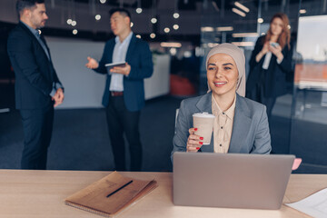 Muslim businesswoman in hijab working on computer and drinking coffee while sitting in modern office