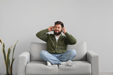 Handsome bearded man in headphones sitting on grey sofa near light wall