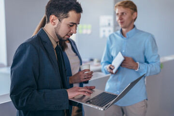Man standing with laptop, his colleagues talking nearby