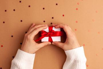 Human hands in white sweater hold a white paper gift with a red satin ribbon bow on brown background with red stars shapes. Christmas Holidays concept flat lay with copy space.