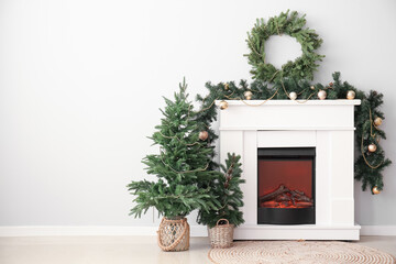 Electric fireplace with Christmas trees, wreath and fir branches near light wall