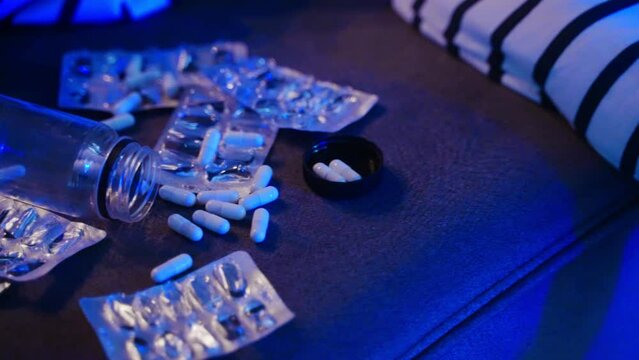 Close Up Of Person Committed Suicide By Drug Overdose Lies On Couch At Home Near Empty Blister Packs, Pills Lit With Red, Blue Police Car, Ambulance Emergency Rotary Lights Drops A Pill From Fingers.