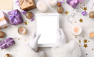 Santa Claus holding tablet computer on white wooden table with Christmas decor and gifts, top view