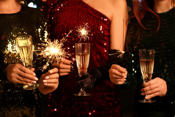 Young women in elegant dresses with burning Christmas sparklers and glasses of champagne, closeup