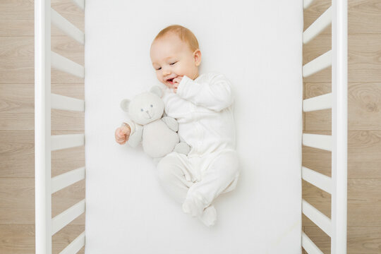 Adorable Happy Baby Boy In White Bodysuit Hugging Teddy Bear And Lying Down On Back On Mattress In Crib At Home Room. 6 Months Old Infant Playing With First Friend. Closeup. Top Down View.