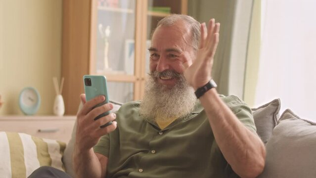 Bearded Caucasian old adult man in wireless earpods video chatting with family on smartphone, sitting on couch in living room at daytime