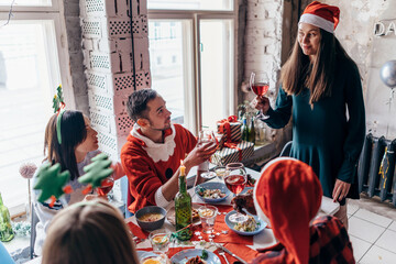 Young woman stands with a glass and says a toast while friends sit at the festive table.