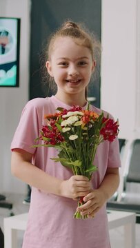 Vertical video: Smiling little girl holding flowers bouquet in hospital waiting area, preparing to visit old patient at health center. Small child sitting in waiting room with mother and father.