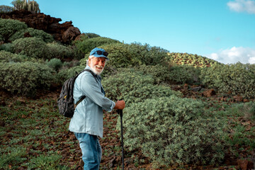 Portrait of old smiling senior man enjoying trekking in countryside. Elderly caucasian male with hat and backpack walking in mountain