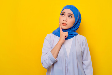 Portrait of pensive young Asian Muslim woman looking up at copy space, thinking about something isolated over yellow background