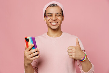 Young gay man wear sweatshirt hat hold in hand use mobile cell phone in rainbow case show thumb up...