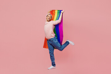 Full size young gay man wear sweatshirt hat hold striped flag behind himself look aside on workspace isolated on plain pastel light pink color background studio portrait Lifestyle lgbtq pride concept