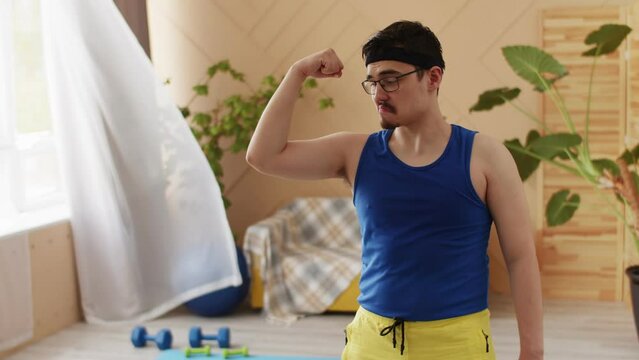 Chubby man with glasses, moustache, goatee in bright blue and yellow retro sportswear showing his arm muscles biceps smiling and nodding positive. Indoors, dumbbells, exercise ball in the background