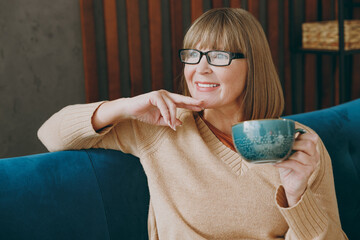 Elderly minded woman 50s year old wear casual clothes sit on blue sofa hold cup drink coffee look...