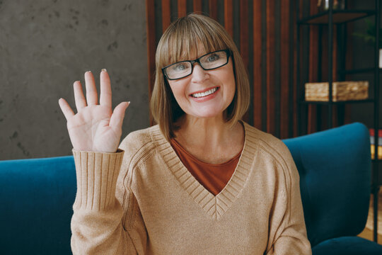 Elderly smiling happy cheerful fun woman 50s year old wears casual clothes sits on blue sofa look camera waving hand stay at home flat rest relax spend free spare time in living room indoor grey wall