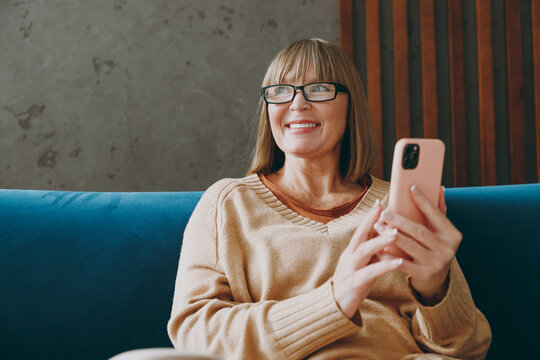 Elderly minded woman 50s year old wear casual clothes sit on blue sofa hold in hand use mobile cell phone look aside stay at home flat rest relax spend free spare time in living room indoor grey wall