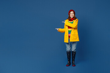 Full body young woman wears sweater red hat yellow waterproof raincoat point index finger aside on workspace isolated on plain dark royal navy blue background Outdoors wet fall weather season concept.