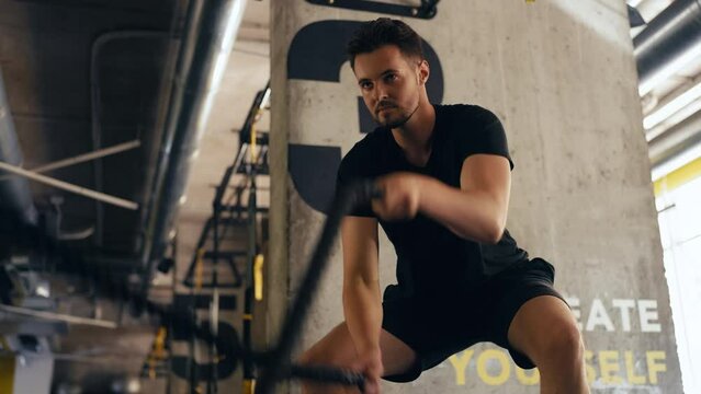 Sporty confident man working out with battle rope in gym, fatburning exercise