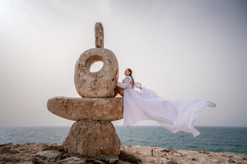 A woman sits on a stone sculpture made of large stones. She is dressed in a white long dress,...