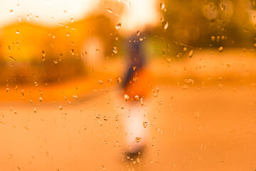 Glass with focused raindrops and blurred background, in an orange environment, with a person in the background. Concept of rain, winter, sadness, dusk, darkness, fear, terror.