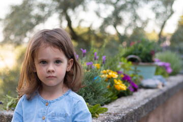 A little beautiful preschooler girl stands in a denim blue dress against the background of flowers