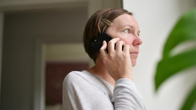 Sad alone woman talking on mobile phone while looking out the window of living room. Casual mid-adult brunette with serious facial expression